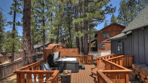 Una terraza de madera con mobiliario exterior, un jacuzzi y una cabaña en un área boscosa.