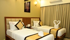 Ein Hotelzimmer mit zwei Betten, jedes mit weißer Bettwäsche und goldfarbenen Akzenten. Darüber hängen Bilder an der Wand, neben den Betten jeweils ein Nachttisch mit Lampe, und am Fenster ein Vorhang.