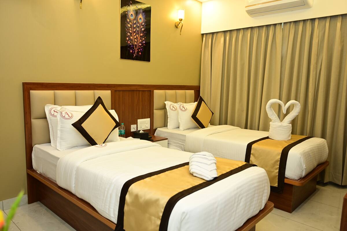 A hotel room with two beds, each with white bedding and a gold accent. There is a wall-mounted artwork above the beds, a nightstand with a lamp, and a curtain on the window.