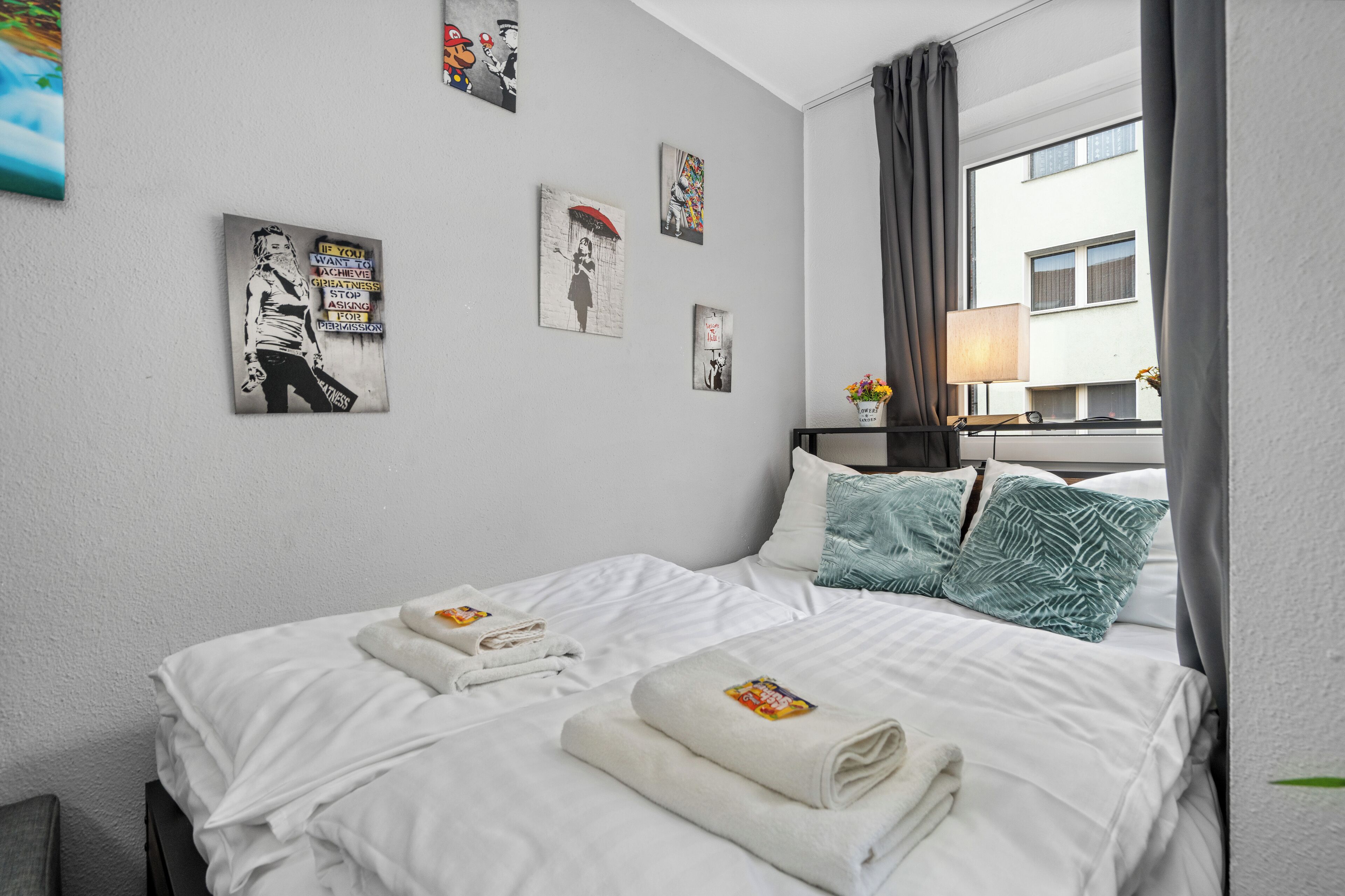 Quarto de hotel com cama, almofadas, mesa de cabeceira, abajur e obras de arte nas paredes.
