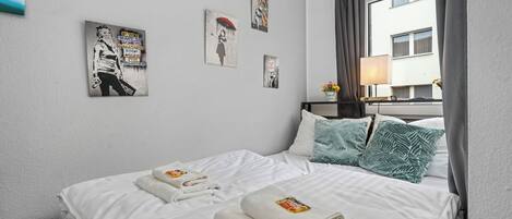 Quarto de hotel com cama, almofadas, mesa de cabeceira, abajur e obras de arte nas paredes.