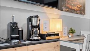 A modern kitchenette with a coffee maker, a kettle, and a microwave. There is a wooden cabinet below the counter and a white chair to the right.