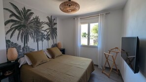 A bedroom with a bed, a wall-mounted TV, a window with curtains, and a wall mural of palm trees. - Beautiful villa with a pool, in a quiet location near Avignon   (Roquemaure)