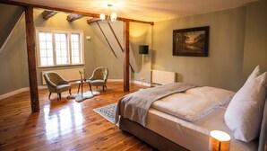 Une chambre avec un grand lit, deux fauteuils, un plafond en bois, un tableau au mur et une fenêtre laissant entrer la lumière naturelle.