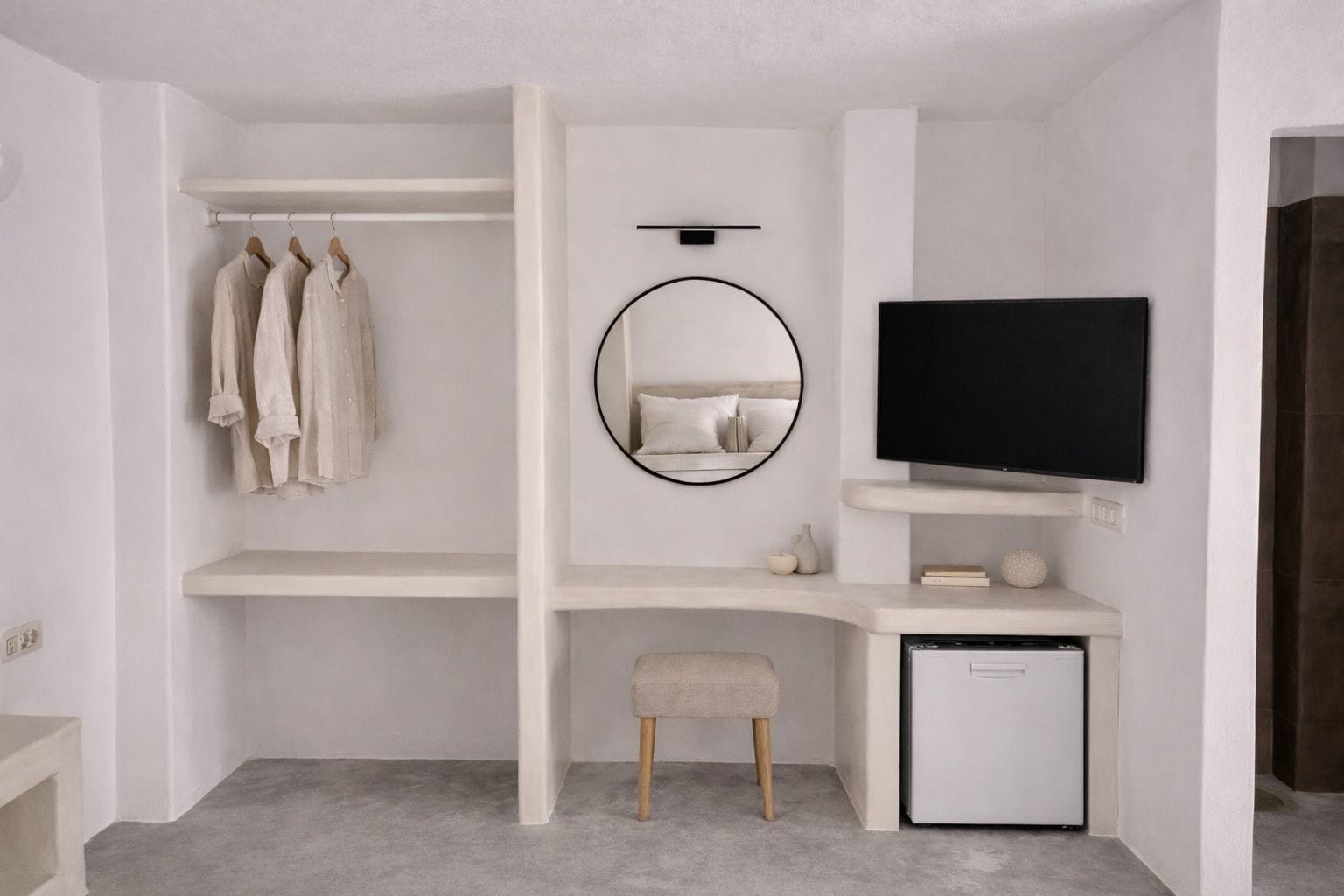 Un dormitorio moderno con escritorio empotrado, televisor, espejo circular y una pequeña zona de estar.