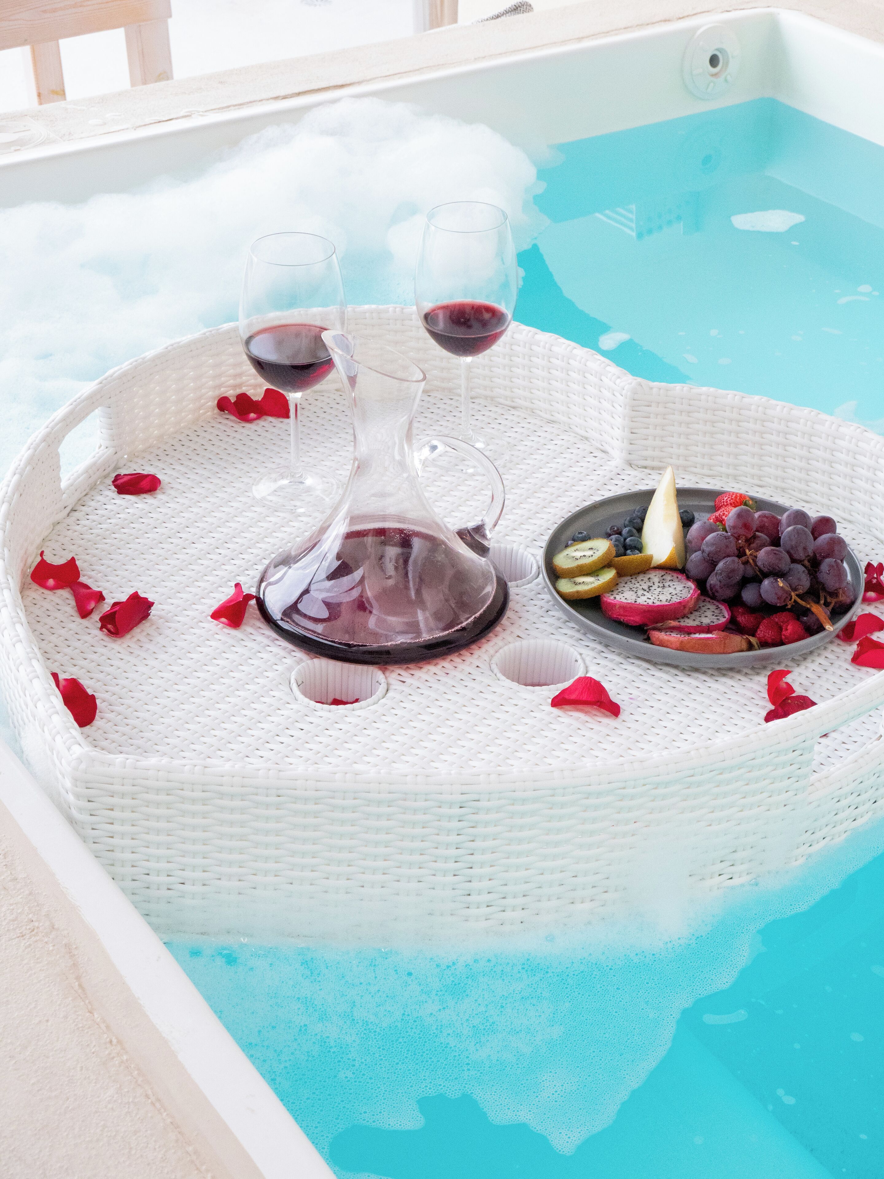 Escena junto a la piscina con una bandeja de mimbre que contiene una botella de vino, dos copas de vino tinto y un cuenco de fruta.