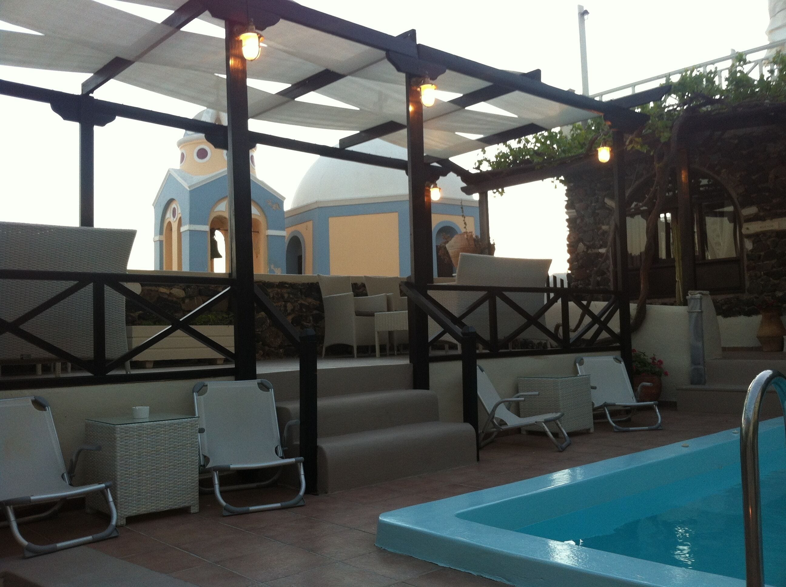 A poolside area with lounge chairs, a covered seating area, and a view of a building with a blue and white facade.
