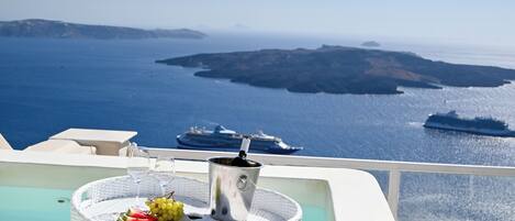 A poolside area with a wicker tray holding a bottle of champagne, two glasses, and a fruit arrangement. In the background, there is a view of the sea and distant islands.