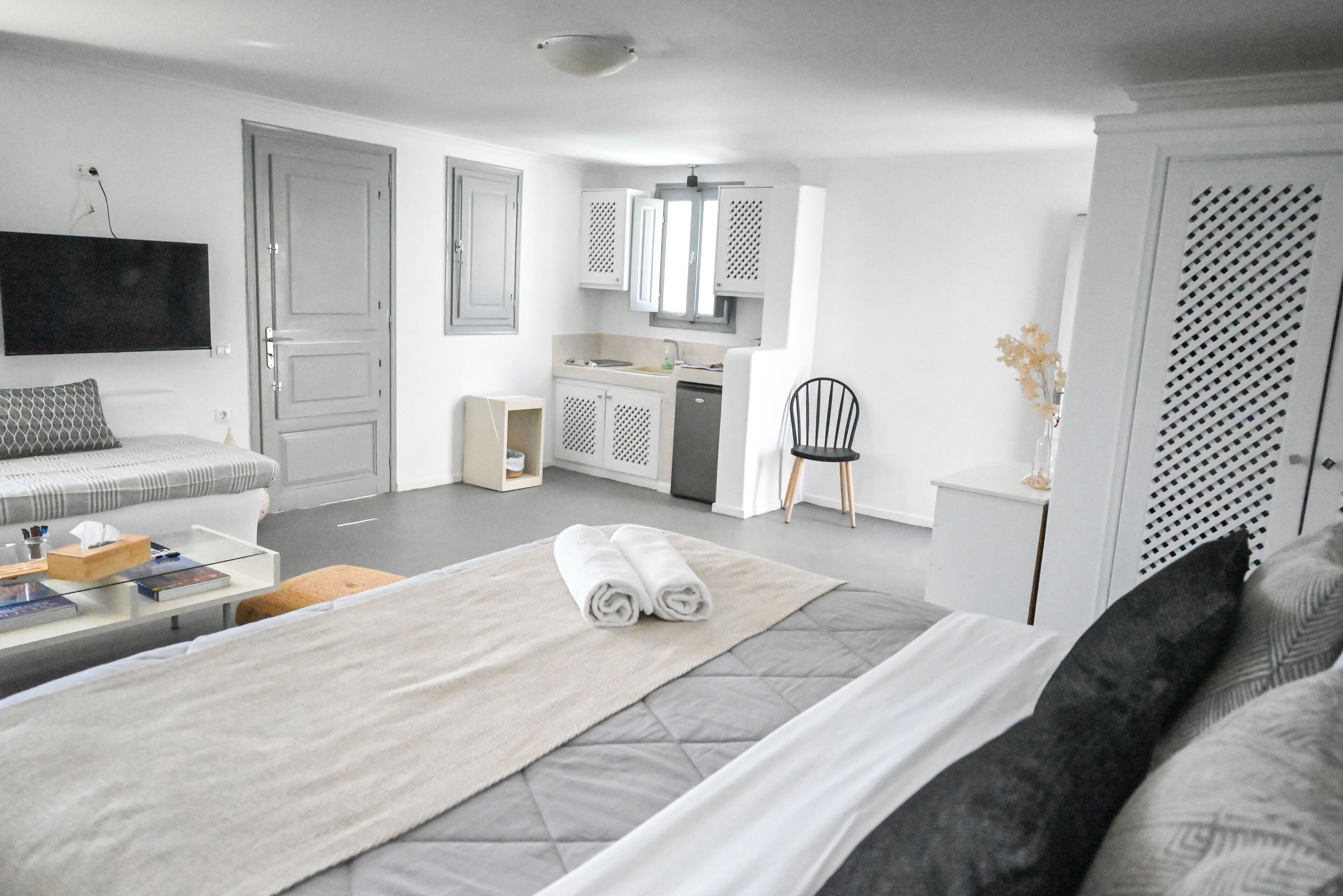 A modern hotel room with a large bed, a sitting area with a sofa and a coffee table, a small kitchen area, and a TV mounted on the wall.