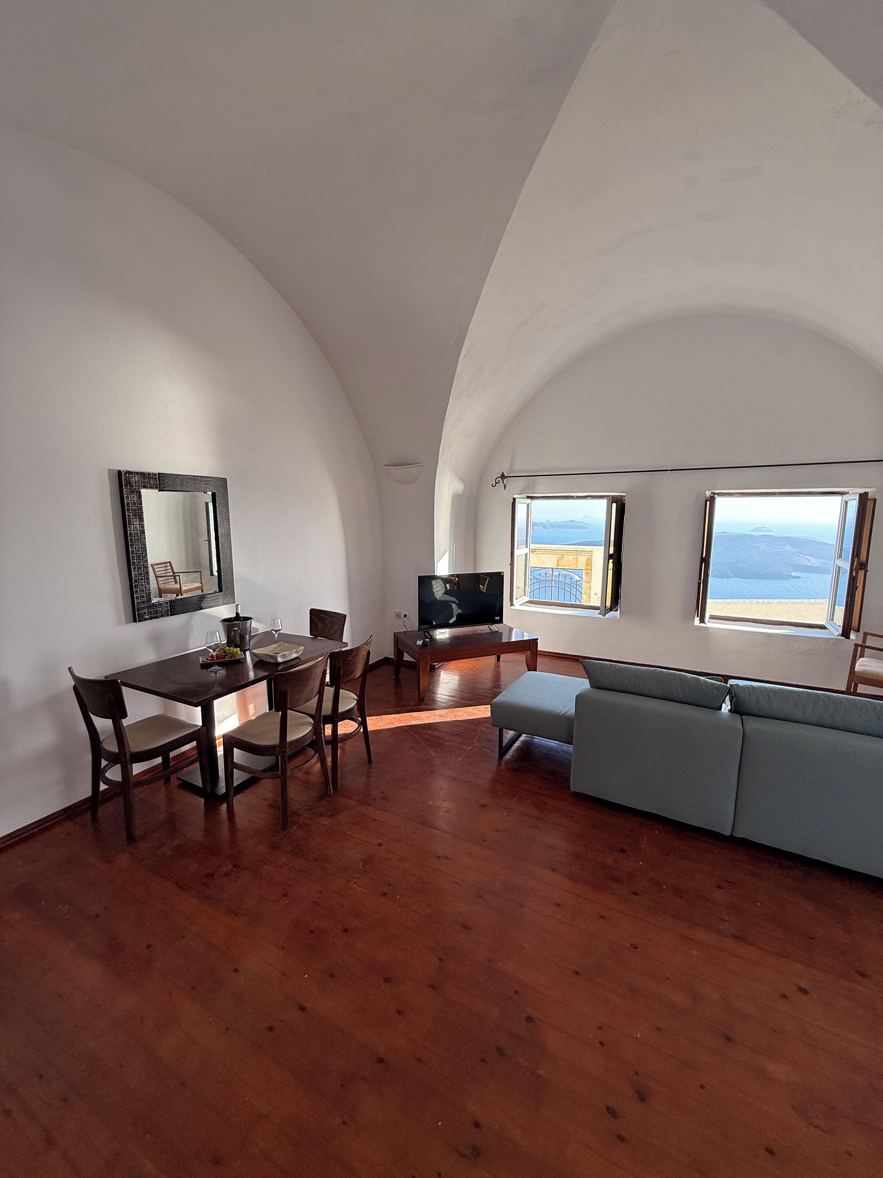 A spacious living area with a dining table, chairs, a sofa, a TV, and large windows offering a view of the sea.