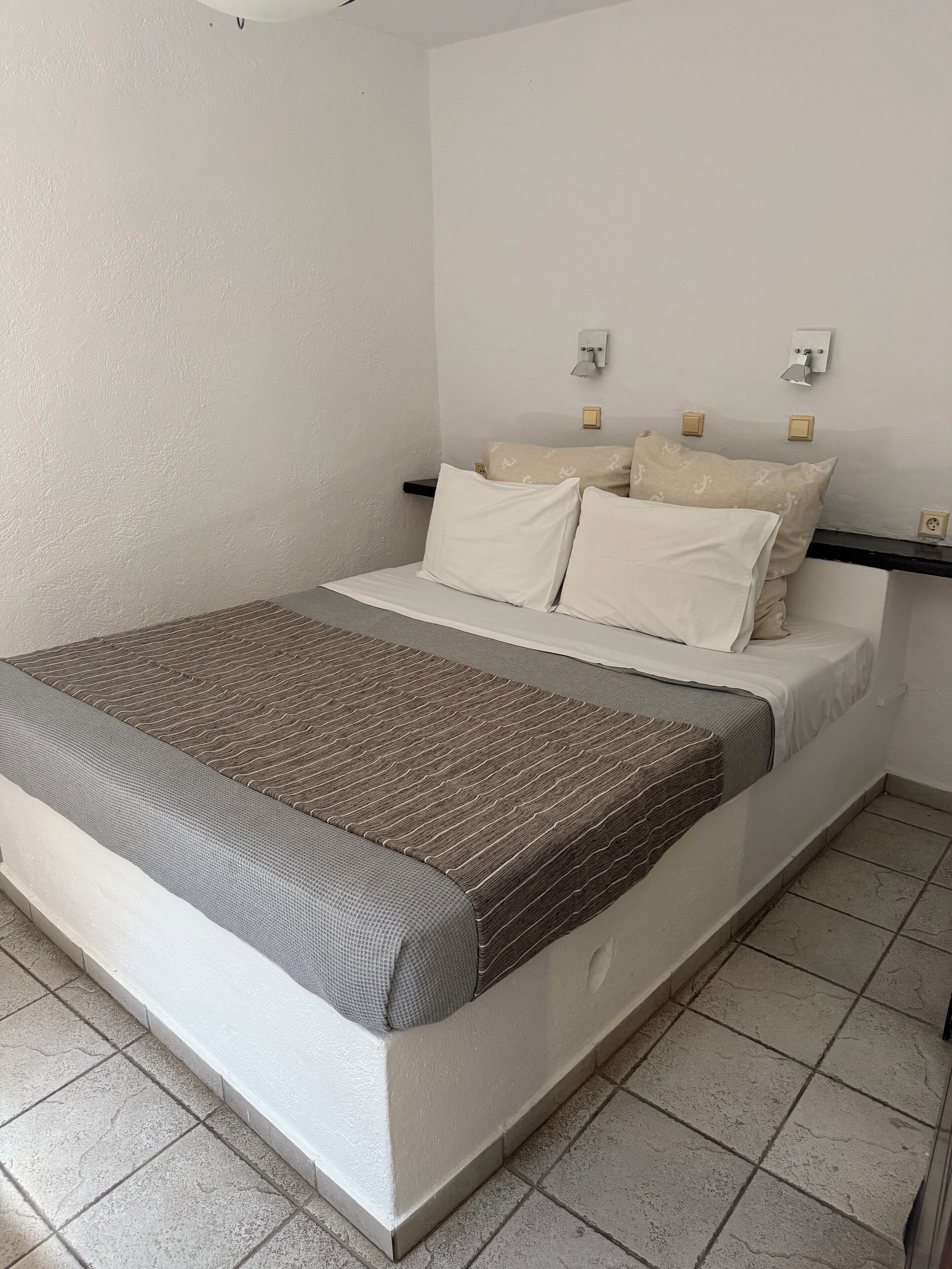 Una cama bien hecha con una colcha a rayas, un cabecero con dos interruptores de luz en la pared y un piso de baldosas.