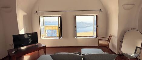 A spacious living room with a large sofa, a television, and two windows offering a view of the sea.