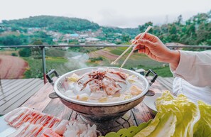 A hot pot meal with various meats and vegetables on a table, set against a scenic outdoor view.