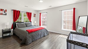 A bedroom with a bed, a nightstand, a window with red curtains, and a view of a brick building outside. - Comfort Oasis - NYC & NWK w/free parking (Newark)