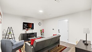 A modern living room with a grey sofa, a small coffee table, a TV mounted on the wall, a ladder, and a door. - Comfort Oasis - NYC & NWK w/free parking (Newark)