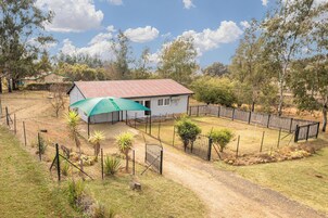 Una casa con techo rojo, rodeada por un patio cercado con poca vegetación y un sendero de tierra que lleva a la entrada.