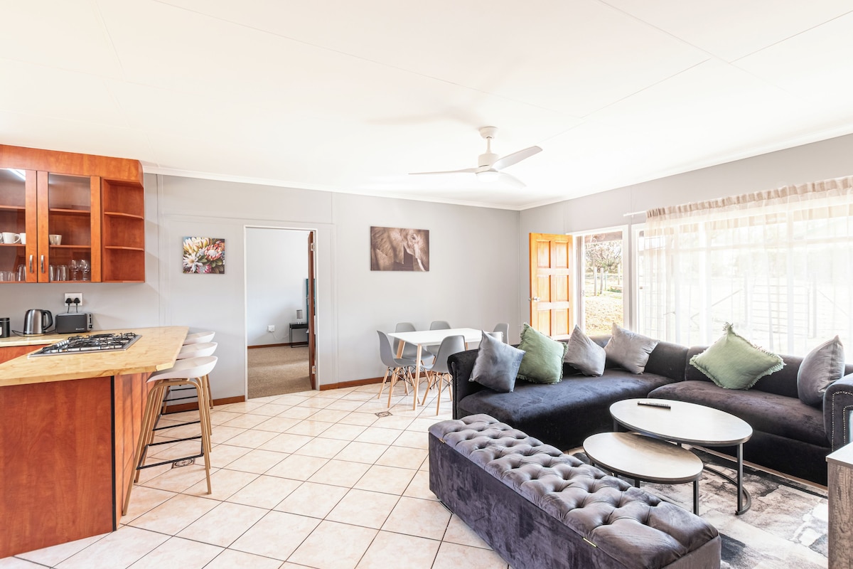 A modern kitchen with a bar area, a dining set, a sofa, and a large window with curtains.