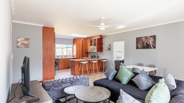 A modern living room with a grey sofa, a round coffee table, and a TV. There is a kitchen area with wooden cabinets and a bar stool. - Forest View Self Catering (Krugersdorp)