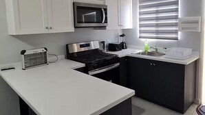 Una cocina compacta con armarios blancos, estufa negra, microondas, pileta y ventana con persianas.