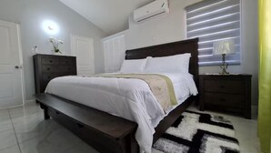 Un dormitorio con cama, mesita de noche, cómoda, aire acondicionado y ventana con persianas.