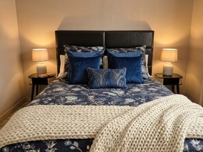 Una cama con una colcha estampada oscura, dos almohadas a juego y un plaid de punto color crema. Hay dos mesitas de noche con lámparas y las paredes son de un tono neutro.