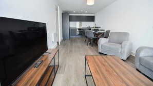 Un salon moderne doté d’un téléviseur à écran plat, d’une table basse en bois, d’un canapé gris et d’un coin repas avec une cuisine en arrière-plan.