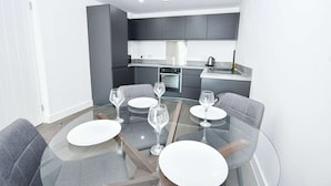 Un espace repas moderne avec une table en verre et des chaises à revêtement gris, une cuisine en arrière-plan avec des meubles sombres et une hotte de cuisine en acier inoxydable.