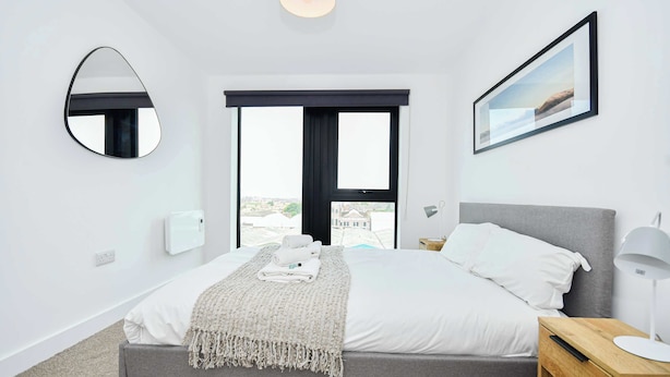 Une chambre à coucher moderne avec un lit, une table de chevet, une commode, un miroir, une fenêtre et un tableau encadré au mur.