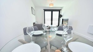 Un coin repas avec une table ronde en verre, des assiettes blanches et des verres à vin. On aperçoit un canapé et un balcon à l’arrière-plan.