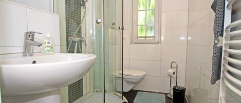Un baño con ducha acristalada, un lavabo blanco, un inodoro y una ventana pequeña.
