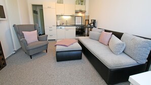 A modern living room with a grey armchair, a grey sofa with cushions, and a black ottoman. A kitchen is visible in the background with white cabinets and a stove. - Modernes Appartement mit Balkon, Wiek (Wiek)