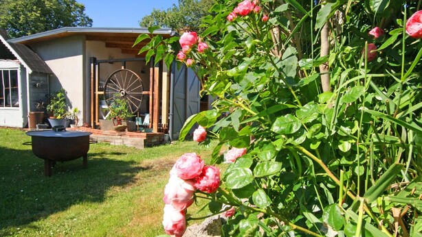 A garden with blooming pink roses, a wooden wheel, and a rustic building with a large window. - Modernes Appartement mit Balkon, Wiek (Wiek)