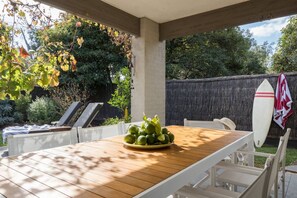 Un patio exterior con una mesa de madera, sillas blancas y una tabla de surf apoyada en la pared.