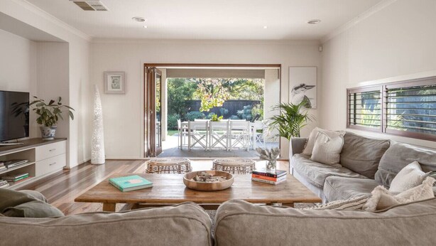 A modern living room with a sofa, coffee table, and TV. There is a dining area with a table and chairs visible through an open door.
