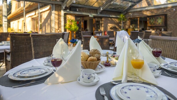 A restaurant setting with tables set for dining, featuring wicker chairs, white tablecloths, and a glass ceiling. - AMAYA Hotel & Suites (Soltau)