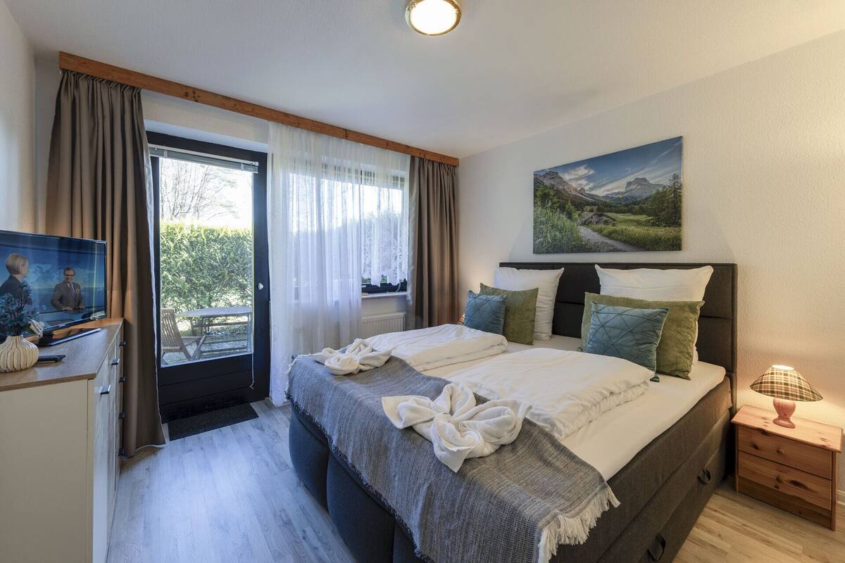 A bedroom with a bed, a bedside table, a lamp, a painting, and a window with curtains.