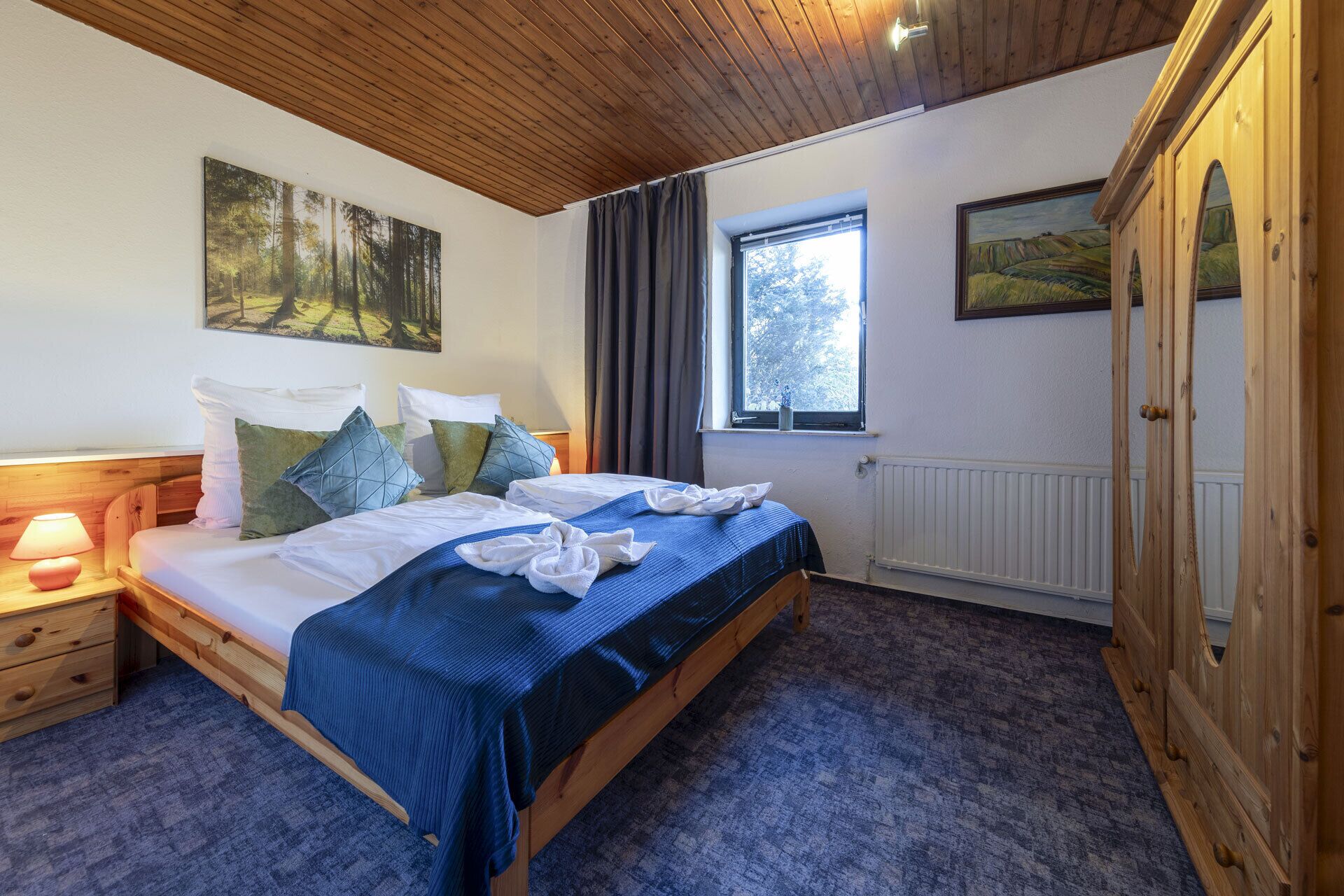 A bedroom with a wooden bed, a nightstand with a lamp, a window with curtains, and a wardrobe.