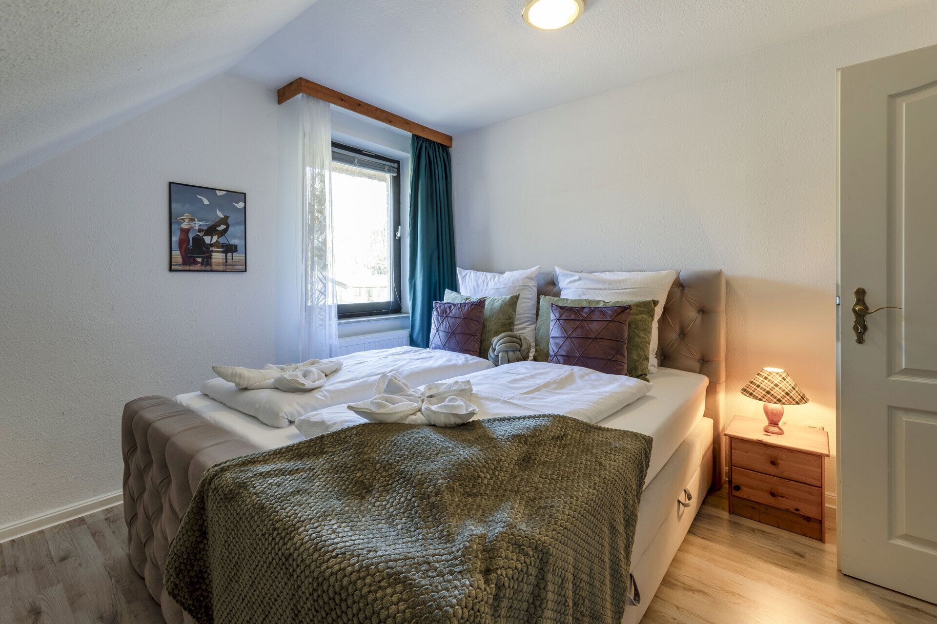 A bedroom with a bed, a window with curtains, a bedside table with a lamp, and a door.