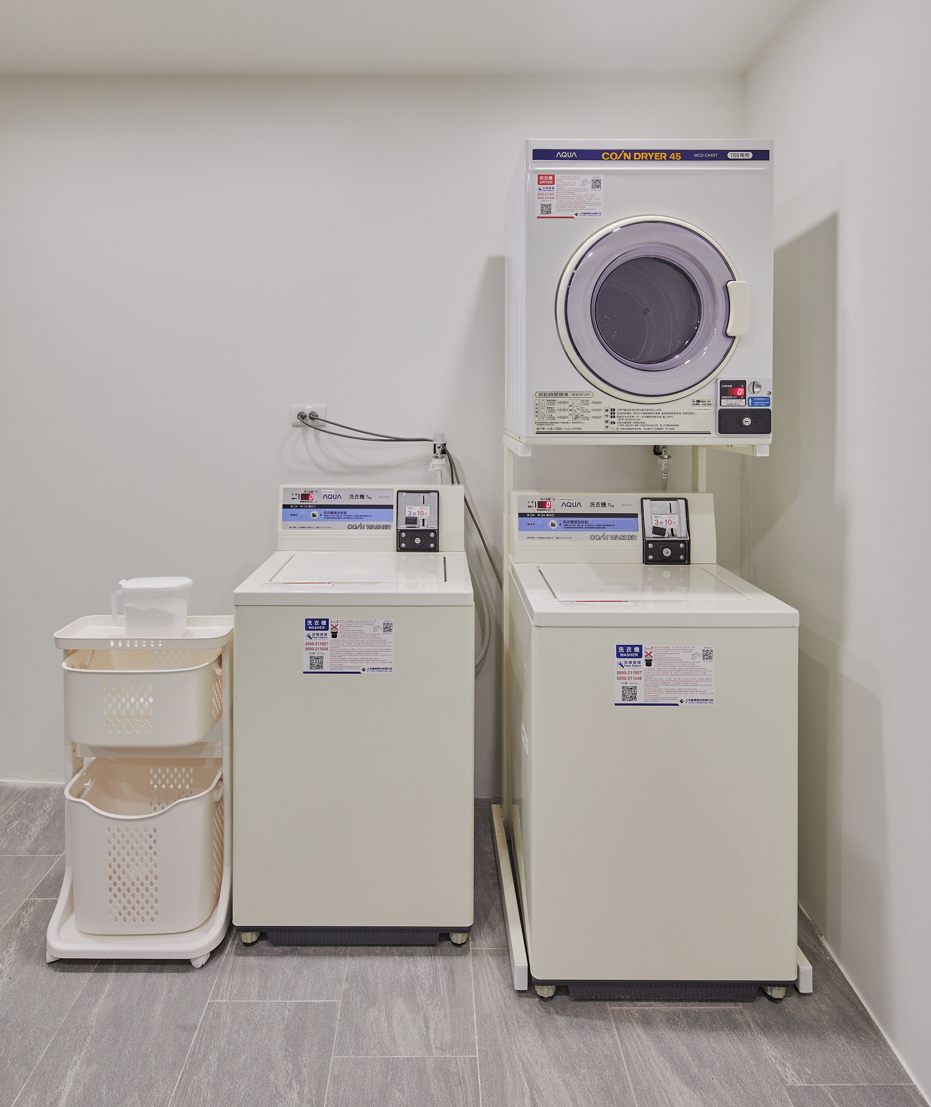 A laundry area with two washing machines and a drying machine, each with control panels and safety labels.