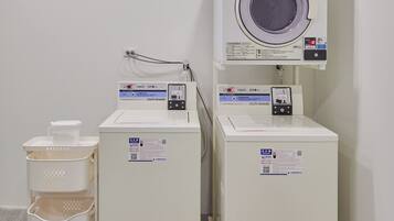 A laundry area with two washing machines and a drying machine, each with control panels and safety labels.