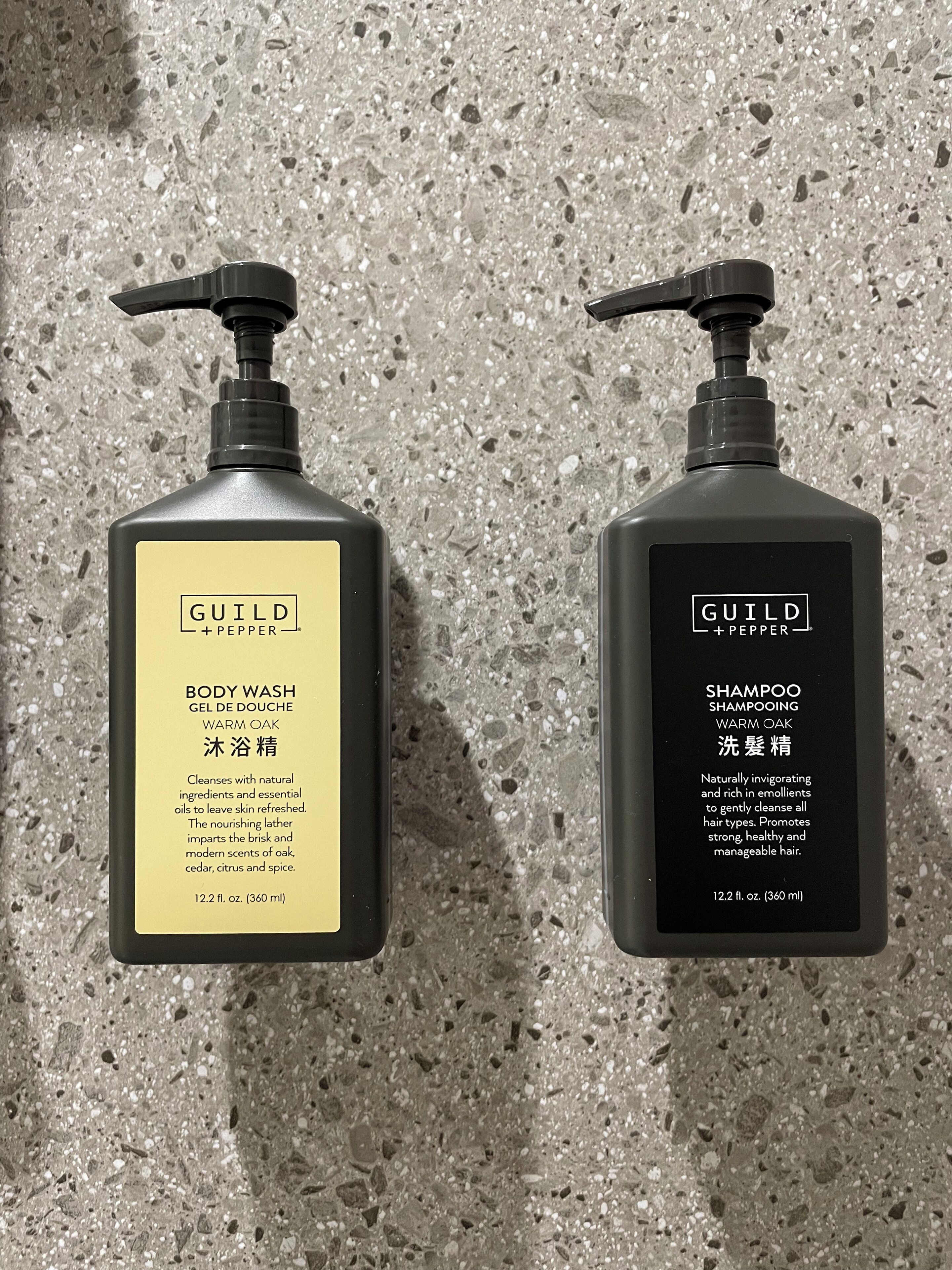 Two bottles of Guild + Pepper body wash and shampoo, each with a pump dispenser, against a speckled concrete background.