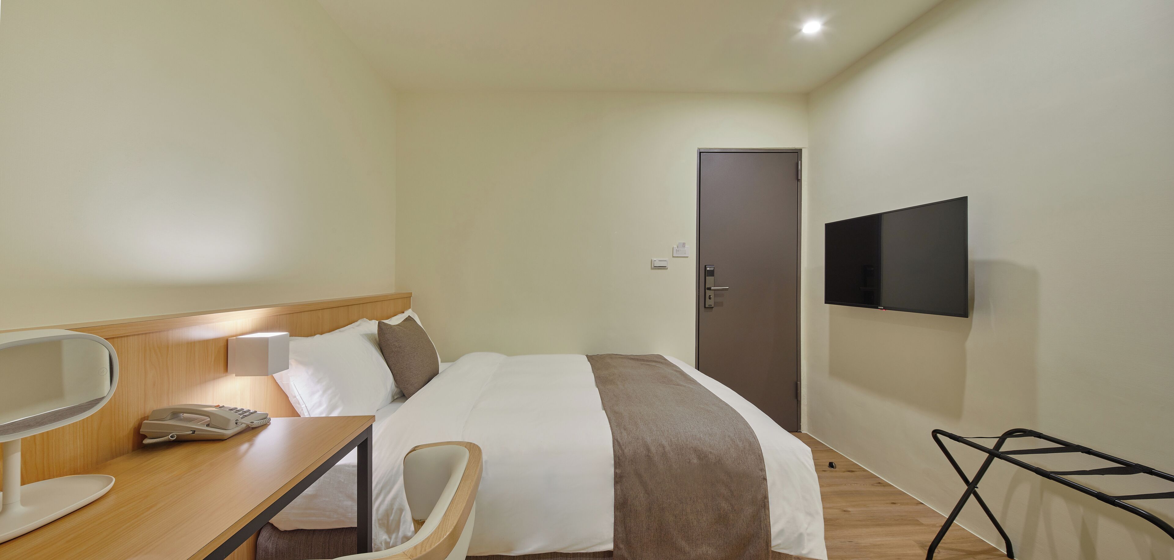 Habitación de hotel con una cama, un escritorio con teléfono, una silla, un televisor montado en la pared y una puerta.