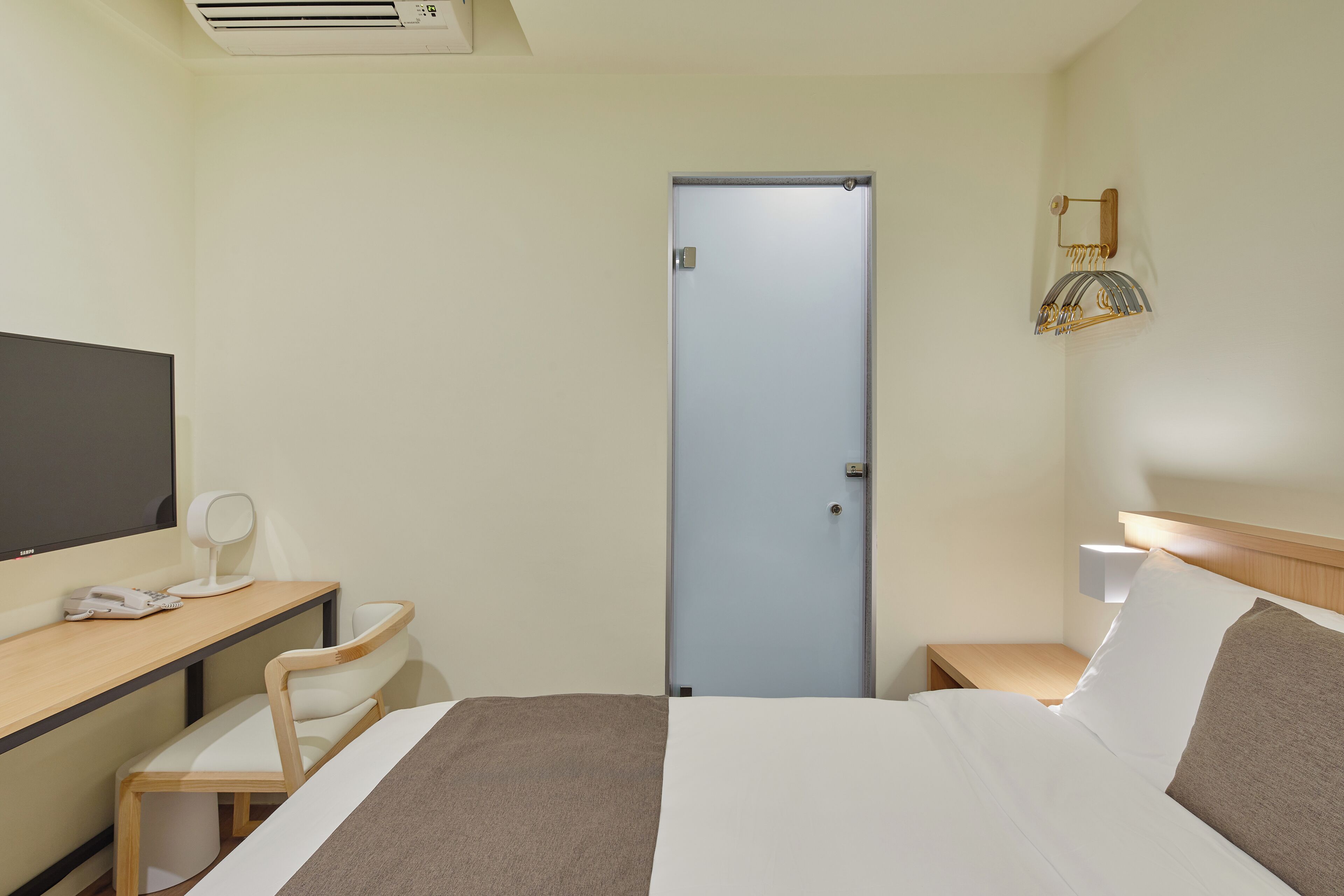 A hotel room with a bed, a desk with a chair, a television, a door, and a wall-mounted lamp.
