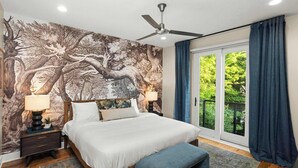 Ein Schlafzimmer mit einem großen Bett, einem Deckenventilator, einem Nachttisch mit Lampe, einer Bank und Blick auf Grün hinter einer Schiebeglastür.
