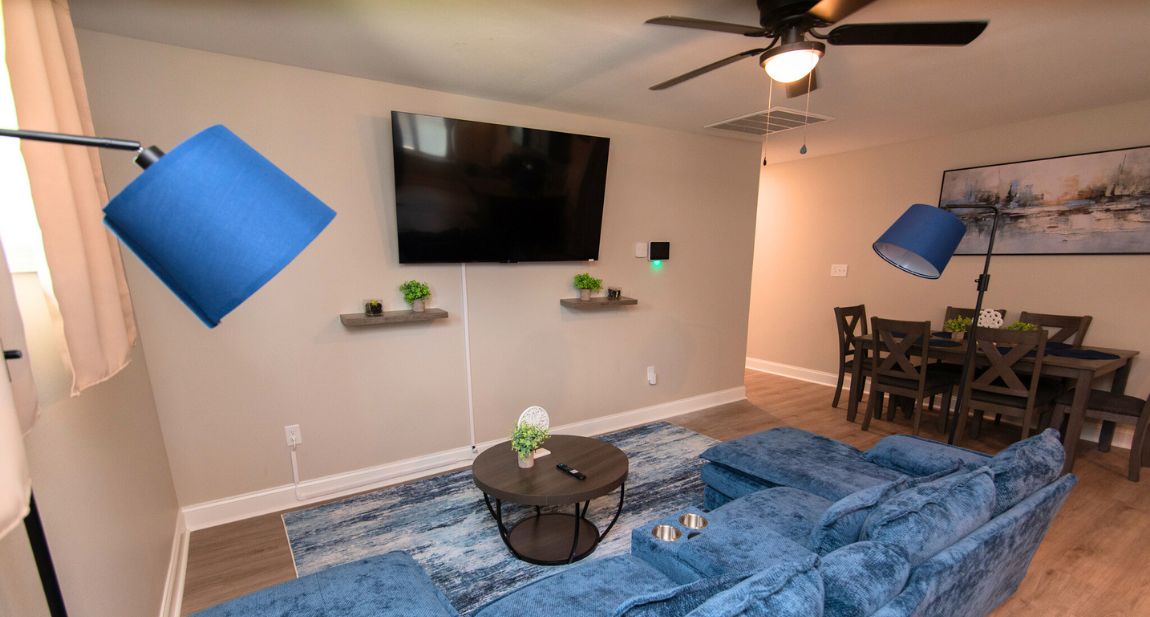 A living room with a large blue sofa, a flat-screen TV, a ceiling fan, and a dining area with a table and chairs.