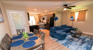 A modern living room with a sectional sofa, a dining table with placemats, a kitchen in the background, and a ceiling fan.