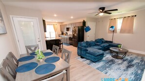 A modern living room with a blue sectional sofa, a dining table with placemats, and a kitchen in the background. - Stylish 3BR Stay w/ Fast Wi-Fi + Parking (Jacksonville)