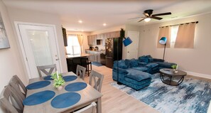 A modern living room with a blue sectional sofa, a dining table with placemats, and a kitchen in the background.