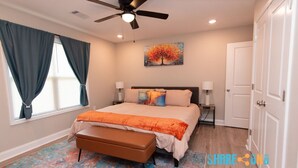 A bedroom with a bed, a ceiling fan, a painting, a bench, and a door.