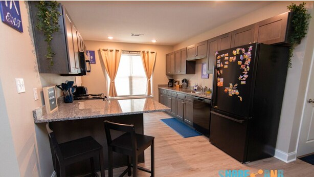 A modern kitchen with granite countertops, dark wooden chairs, a black refrigerator, and a window with curtains.